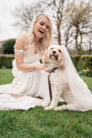 A fluffy white dog sits with someone in a wedding dress on green grass during an outdoor photo session.