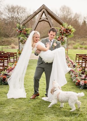 A wedding couple plays with their fluffy white dog in a grassy outdoor ceremony setting with wooden chairs.