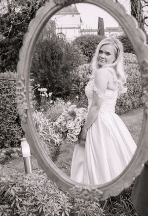 Black and white reflection in ornate mirror shows a bride in a classic white wedding dress posing in a garden setting.