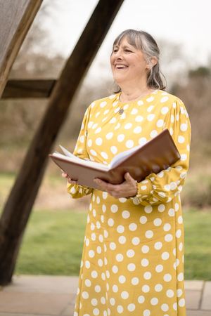 A person in a bright yellow polka dot dress reads from a book while standing outdoors on a sunny day.