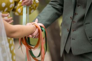 A couple holds hands during a wedding ceremony while exchanging colorful ribbons in a traditional handfasting ritual.