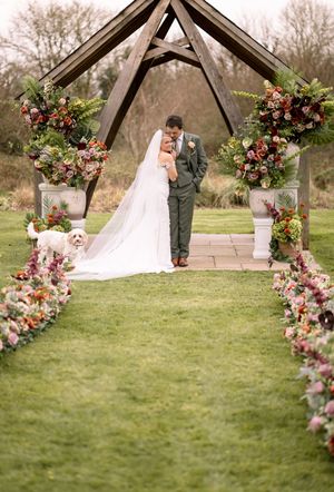 A romantic outdoor wedding ceremony under a wooden arch decorated with lush pink and burgundy floral arrangements on a green lawn.