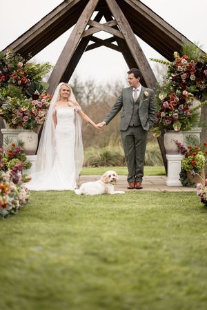 Wedding couple holds hands under a wooden gazebo decorated with burgundy and pink floral arrangements on a grassy lawn.