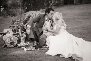 A couple in wedding attire relaxes on a blanket during an outdoor picnic celebration.
