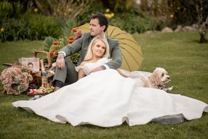 A couple in wedding attire relaxes on a blanket in a lush green garden setting during their outdoor celebration.
