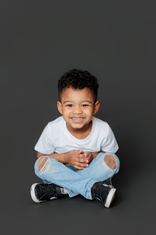 A child wearing a white t-shirt and light blue pants sits cross-legged on a dark studio background.