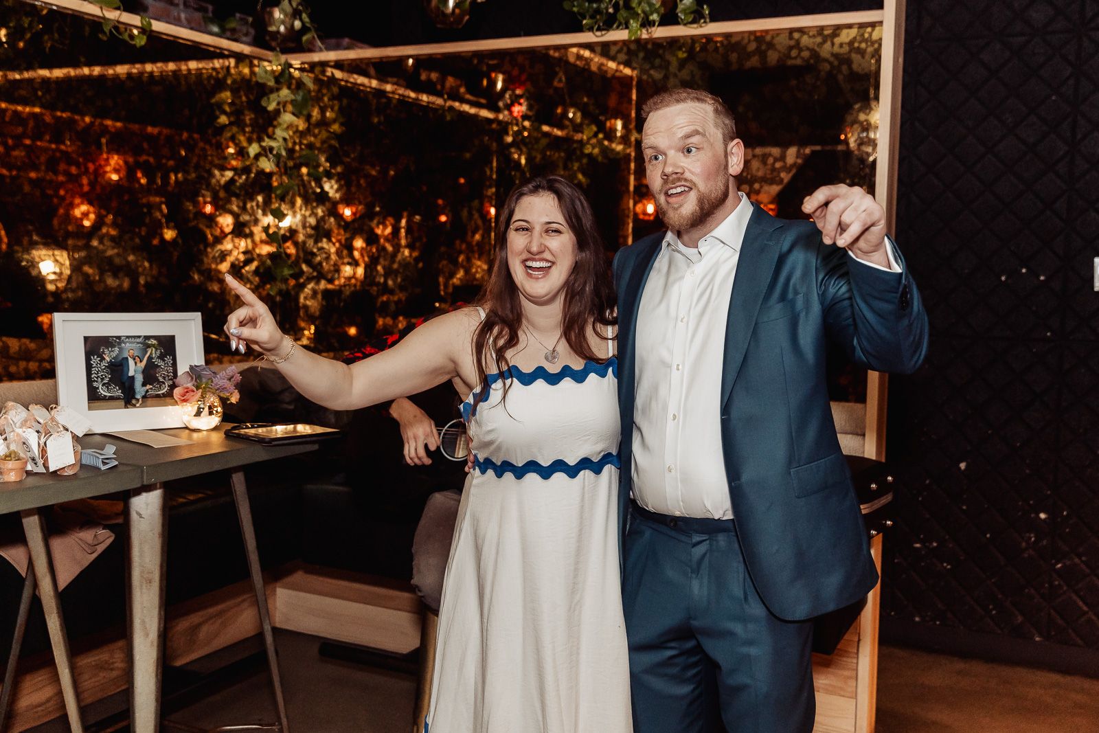 Couple in formal attire smiling and pointing at a framed photo at an elegant evening event.