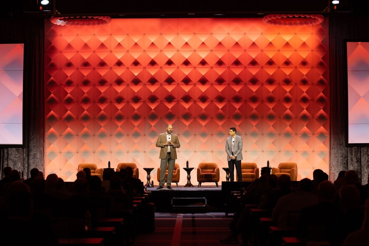 Two speakers stand on a stage with a bold red geometric wall backdrop and orange chairs at a conference event in Dallas, TX