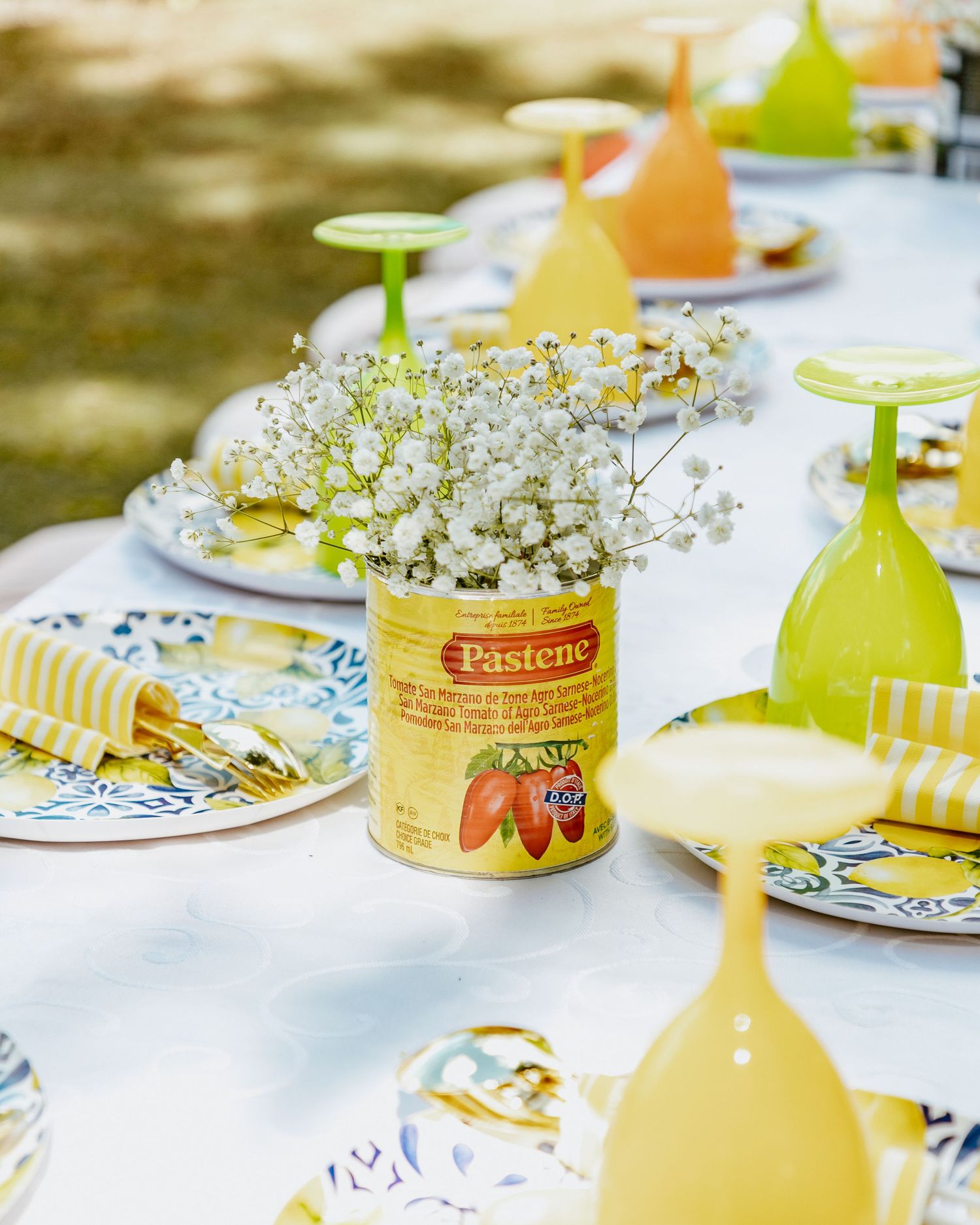 Outdoor table setting with Pastene tomato can vase holding baby's breath, yellow glassware, and blue patterned plates.