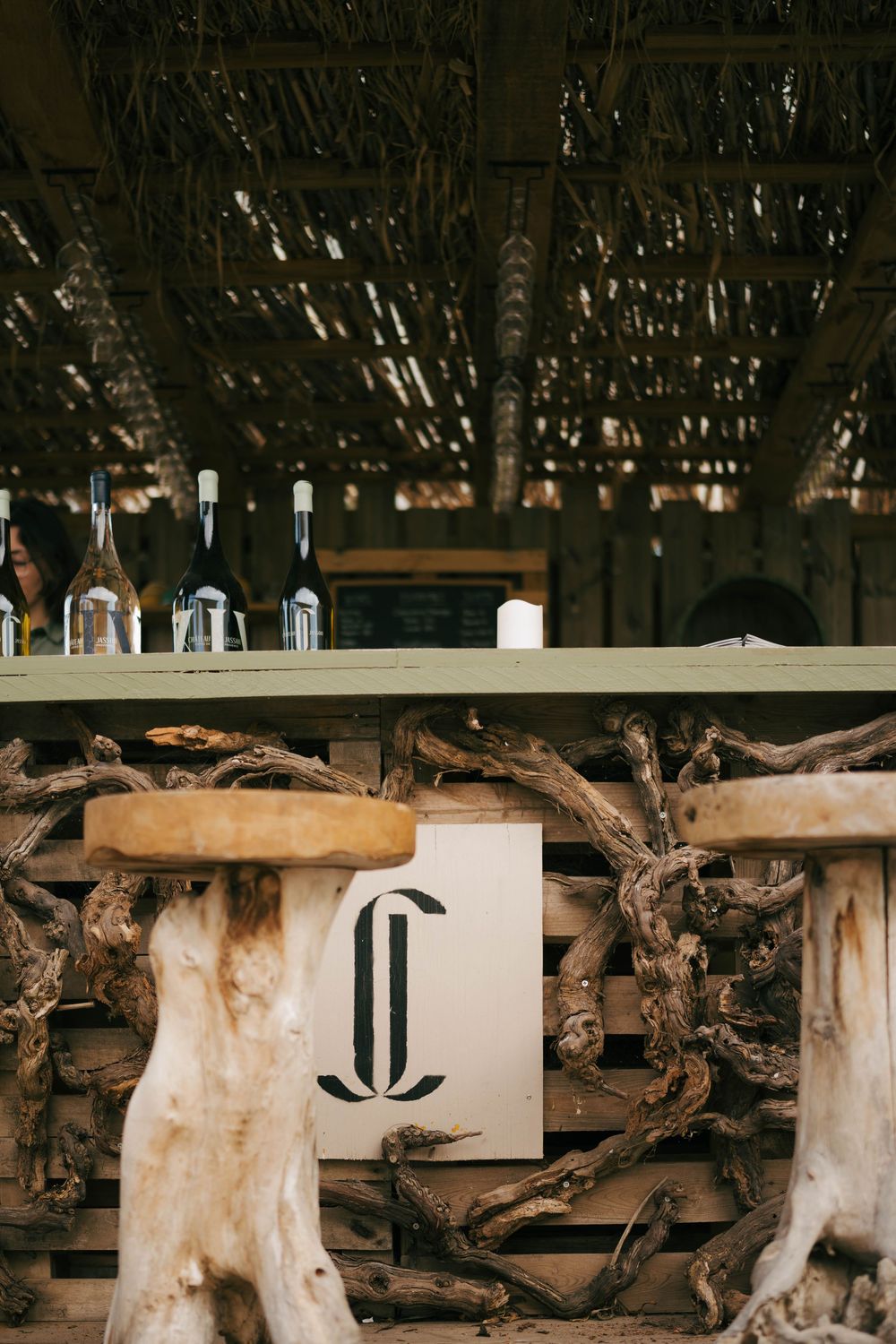 Rustic outdoor bar with driftwood stools, vine-wrapped counter, bottles on shelf, and thatched roof canopy.