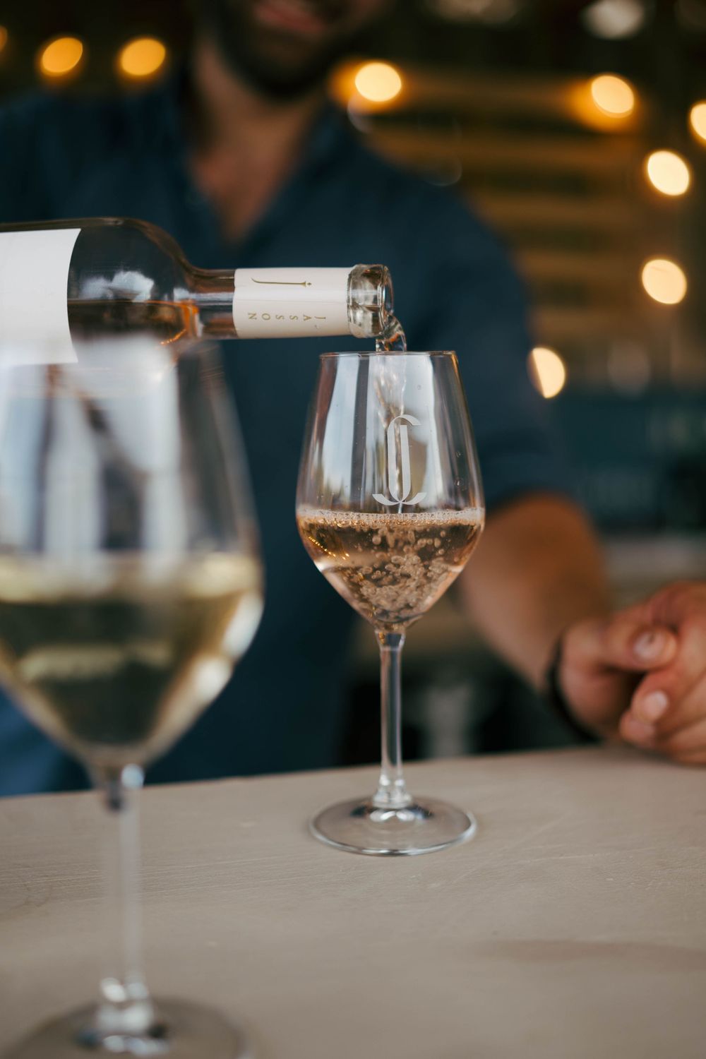 Un homme en chemise bleue versant du vin blanc dans un verre à une table de restaurant avec des lumières chaudes de bokeh.