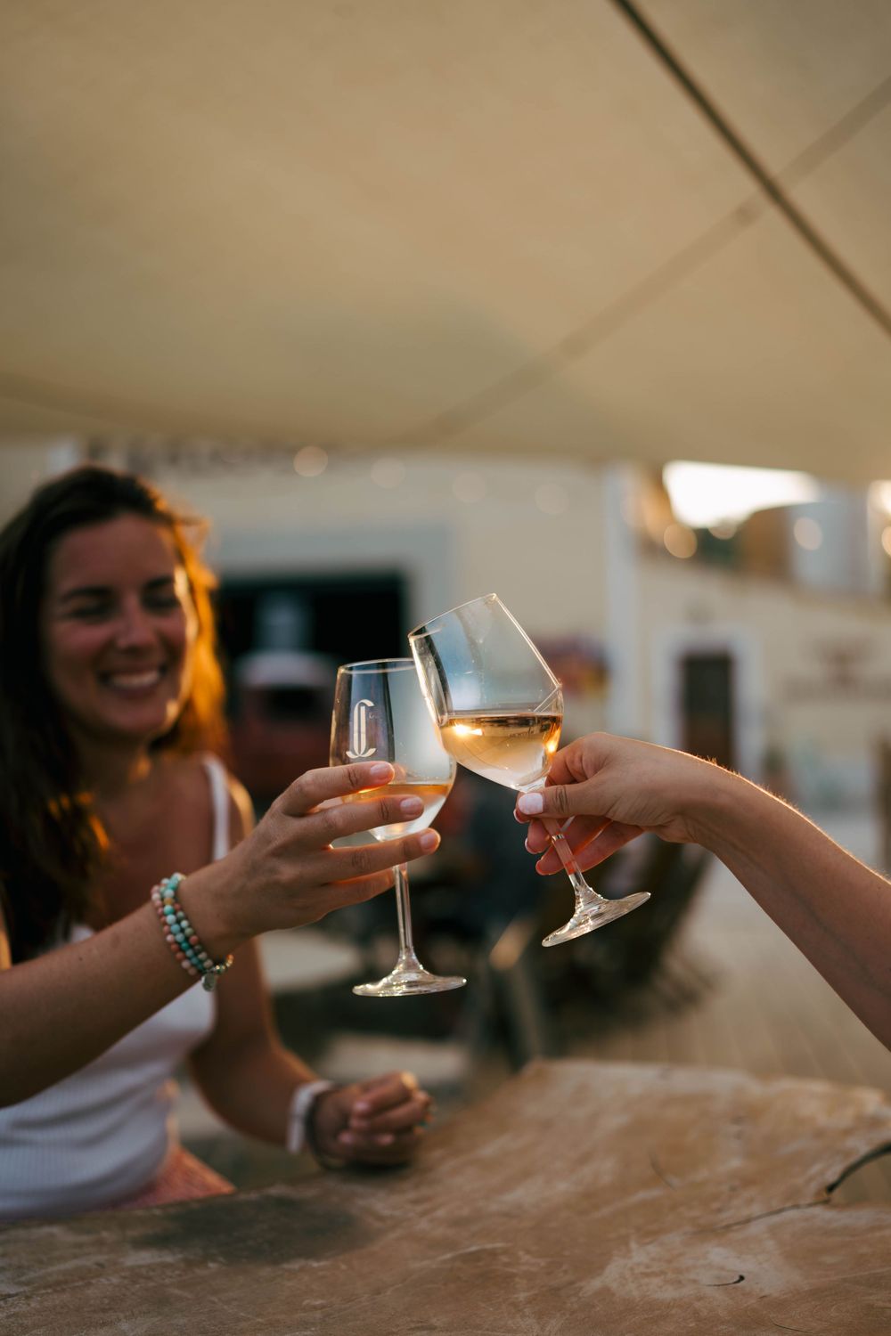 Two women toasting with white wine glasses at an outdoor bar during golden hour, smiling and celebrating together.