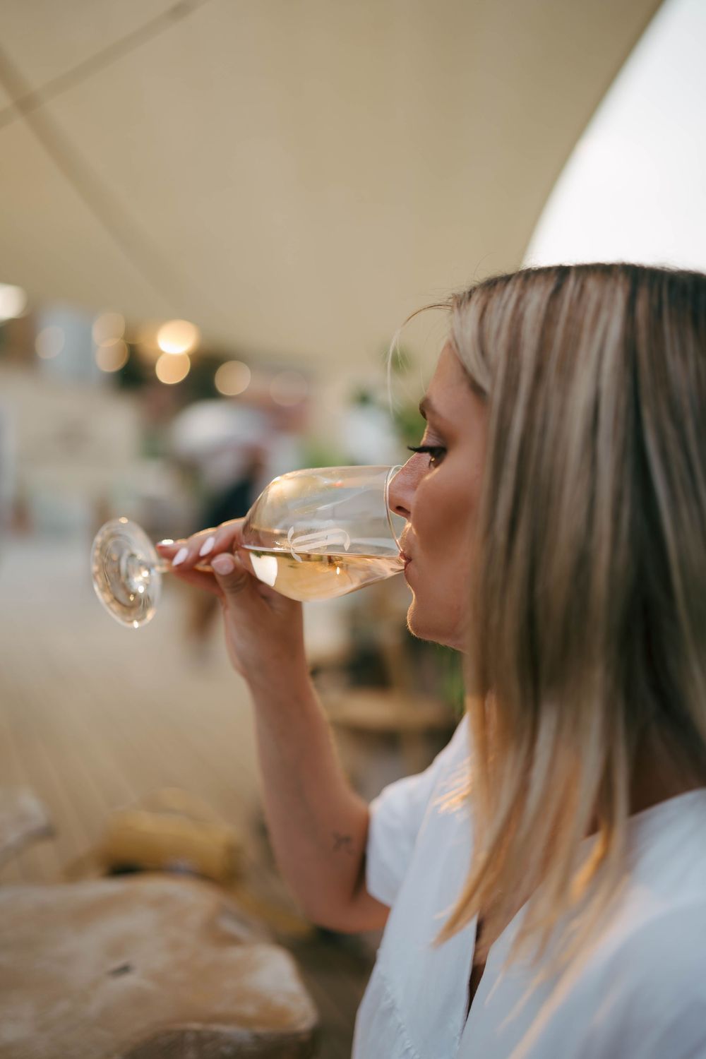 Femme blonde sirotant du vin blanc dans un verre lors d'un événement en plein air sous une tente.