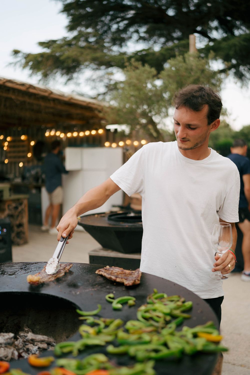 Homme en t-shirt blanc faisant griller de la viande et des légumes sur un barbecue en plein air tout en tenant un verre de vin au crépuscule