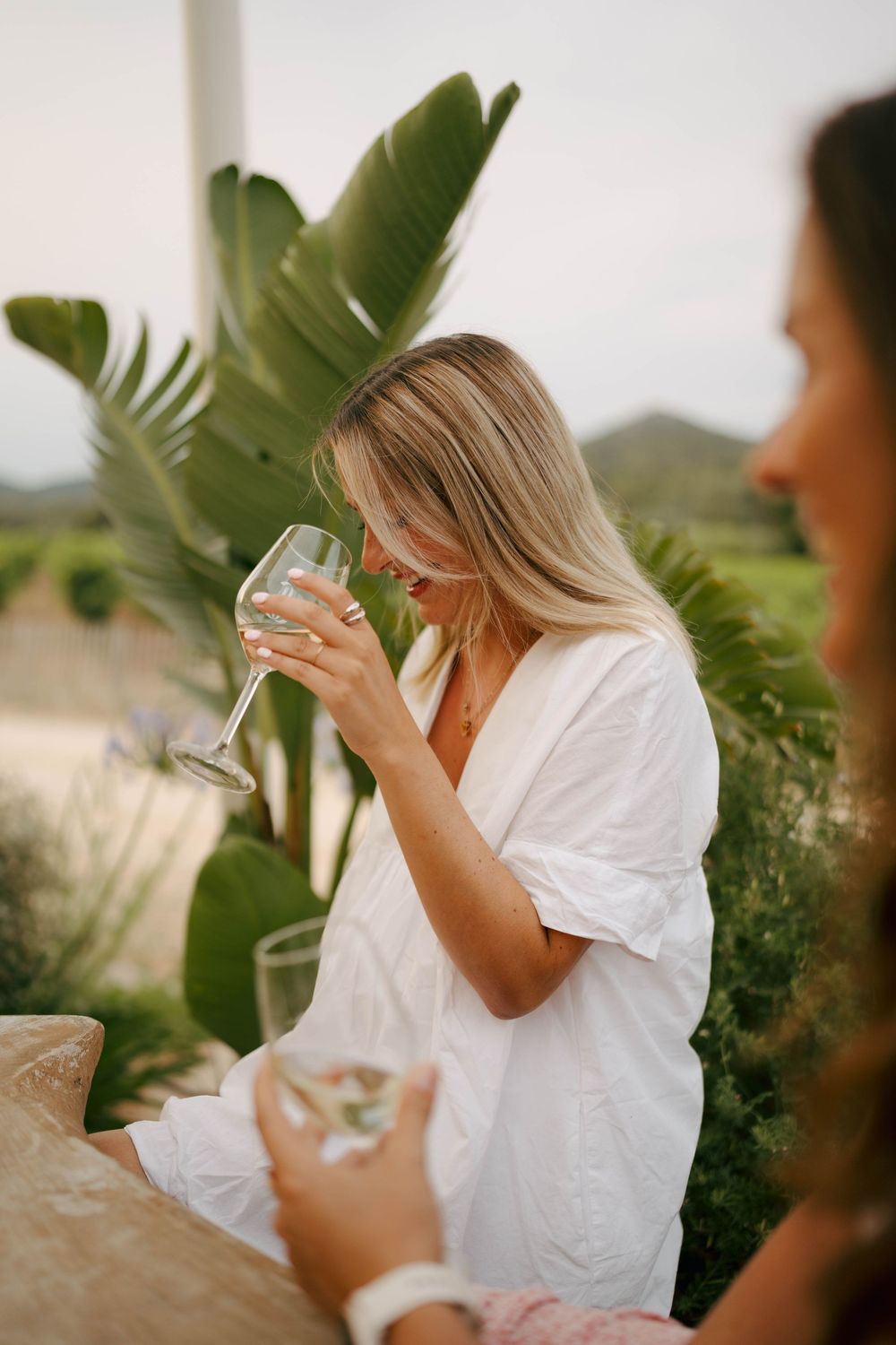 Femme en robe blanche sirotant du vin blanc à l'extérieur, avec des plantes tropicales et des vues panoramiques sur les vignobles en arrière