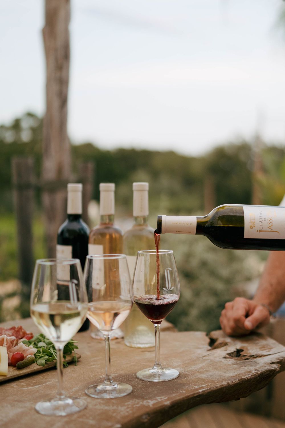 Du vin rouge versé dans un verre à une table de dégustation de vin en plein air avec des vins blancs et une planche de charcuterie.