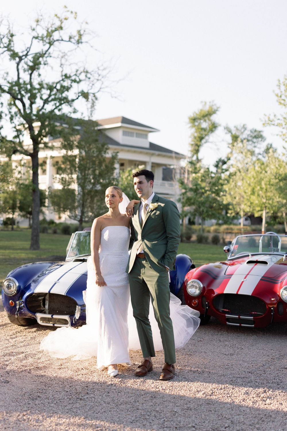 Couple in wedding attire pose between two classic vintage Shelby Cobra sports cars on a gravel driveway.