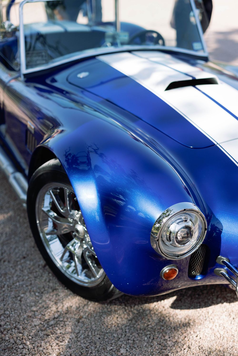 Close up of a classic blue Shelby Cobra sports car's front fender showing chrome headlight and white racing stripes.