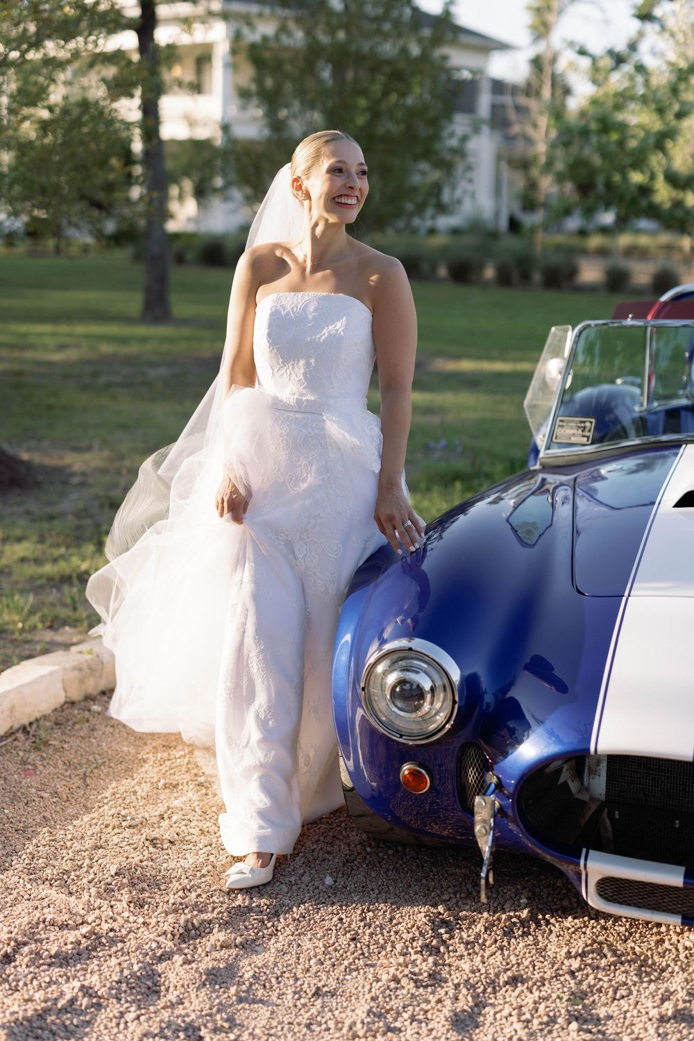 A bride in a white wedding dress poses next to a vintage blue and white classic car on a sunny day.