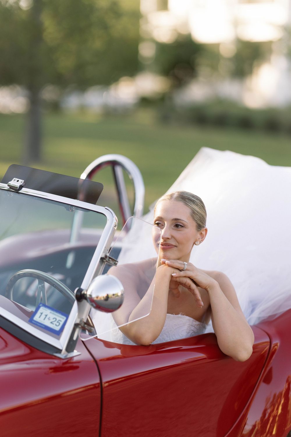 A vintage red convertible car sits parked in a sunny garden setting with a bride in an elegant white dress.