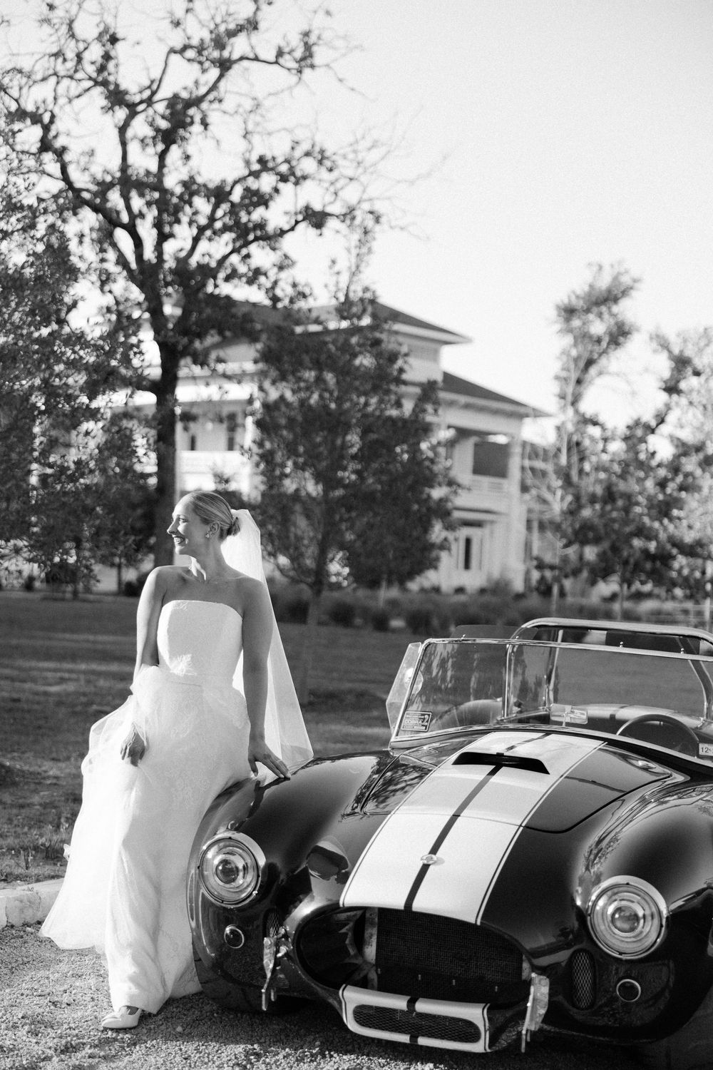 Black and white photo of a bride in white dress posing with a vintage Shelby Cobra convertible on a mansion estate lawn.