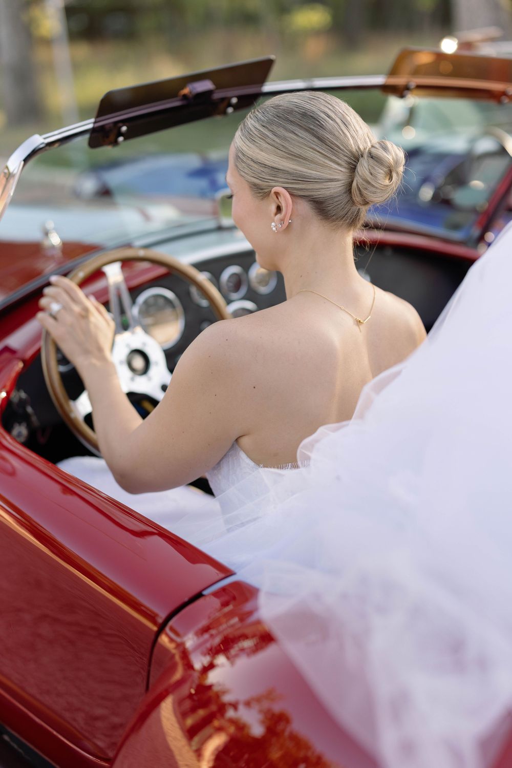 Person wearing white wedding dress sits in driver's seat of classic red convertible car on sunny day.