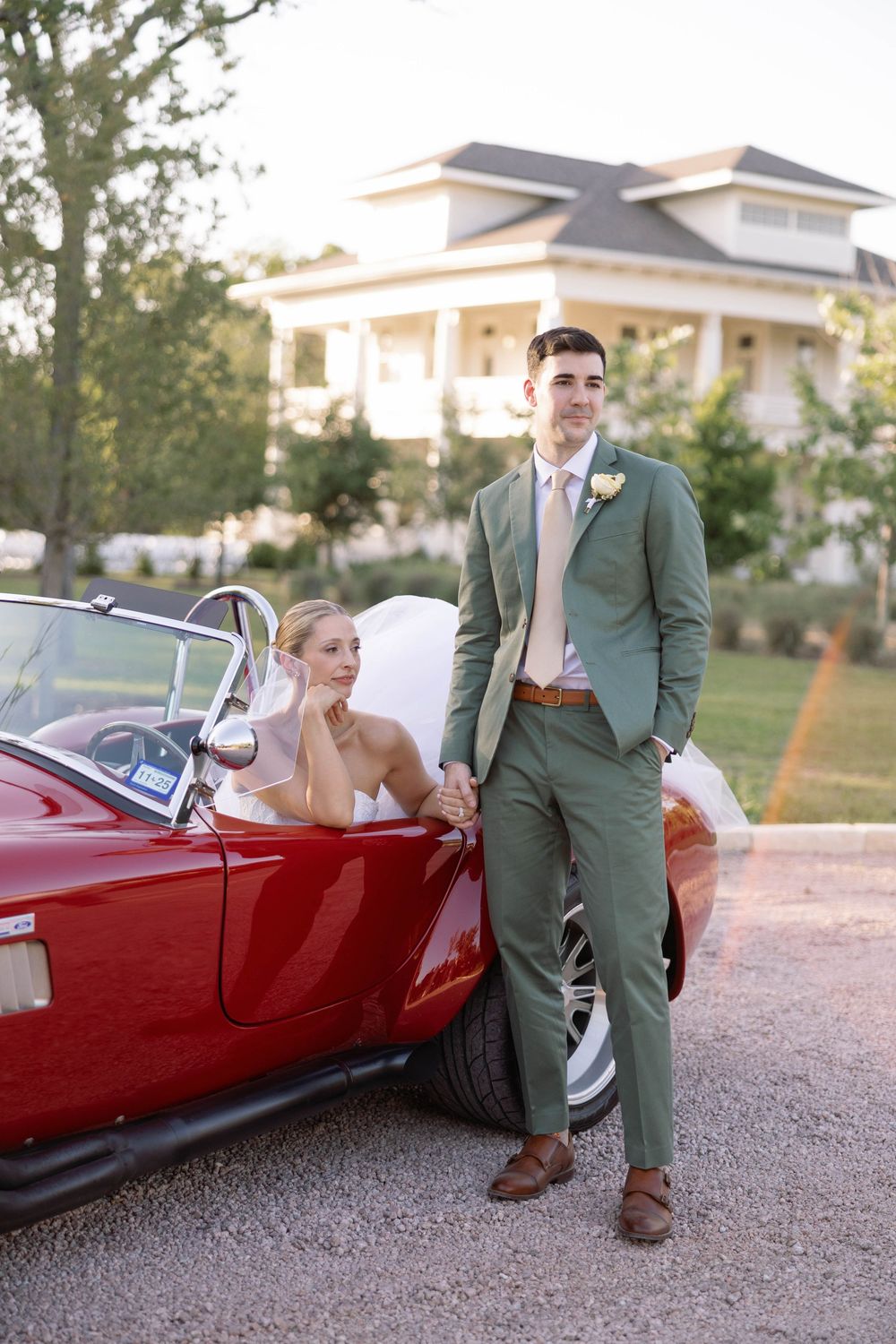 A couple poses with a classic red vintage car outside a beautiful white estate house at sunset.