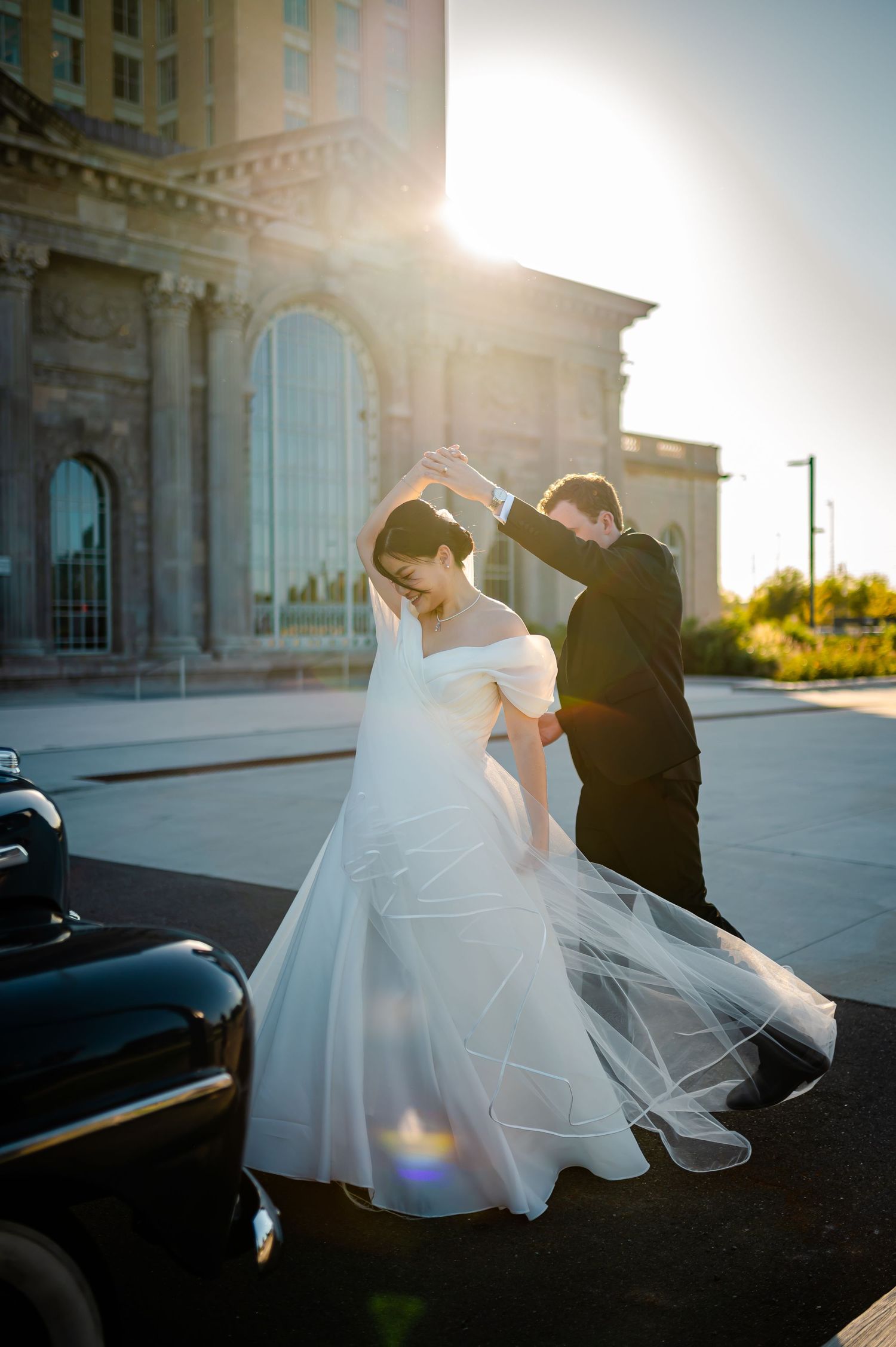 A couple in wedding attire dances together near a vintage car as golden sunlight streams through The Michigan Central Station