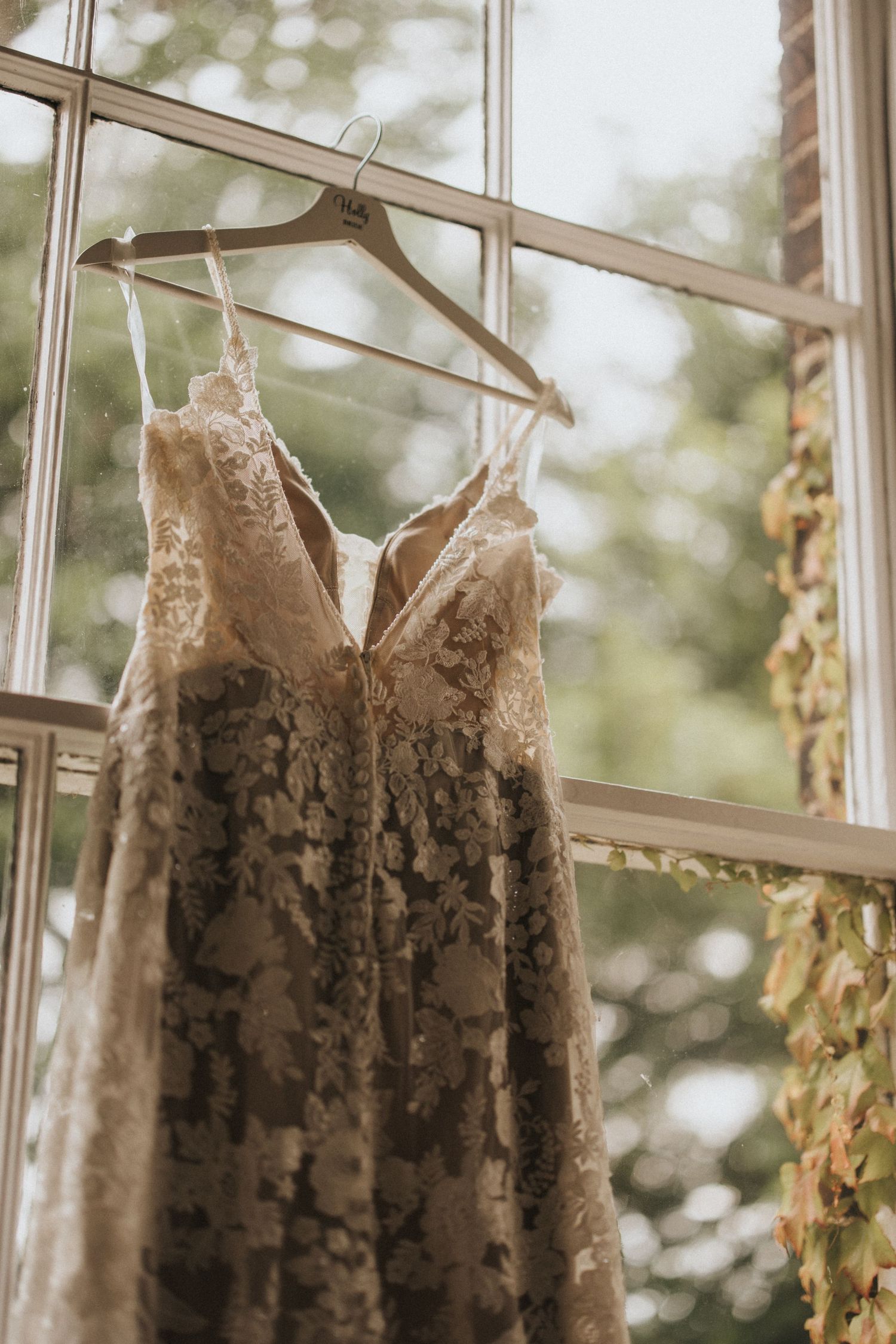 A delicate white lace wedding dress hangs in front of a sunlit window with beige curtains in an elegant room.