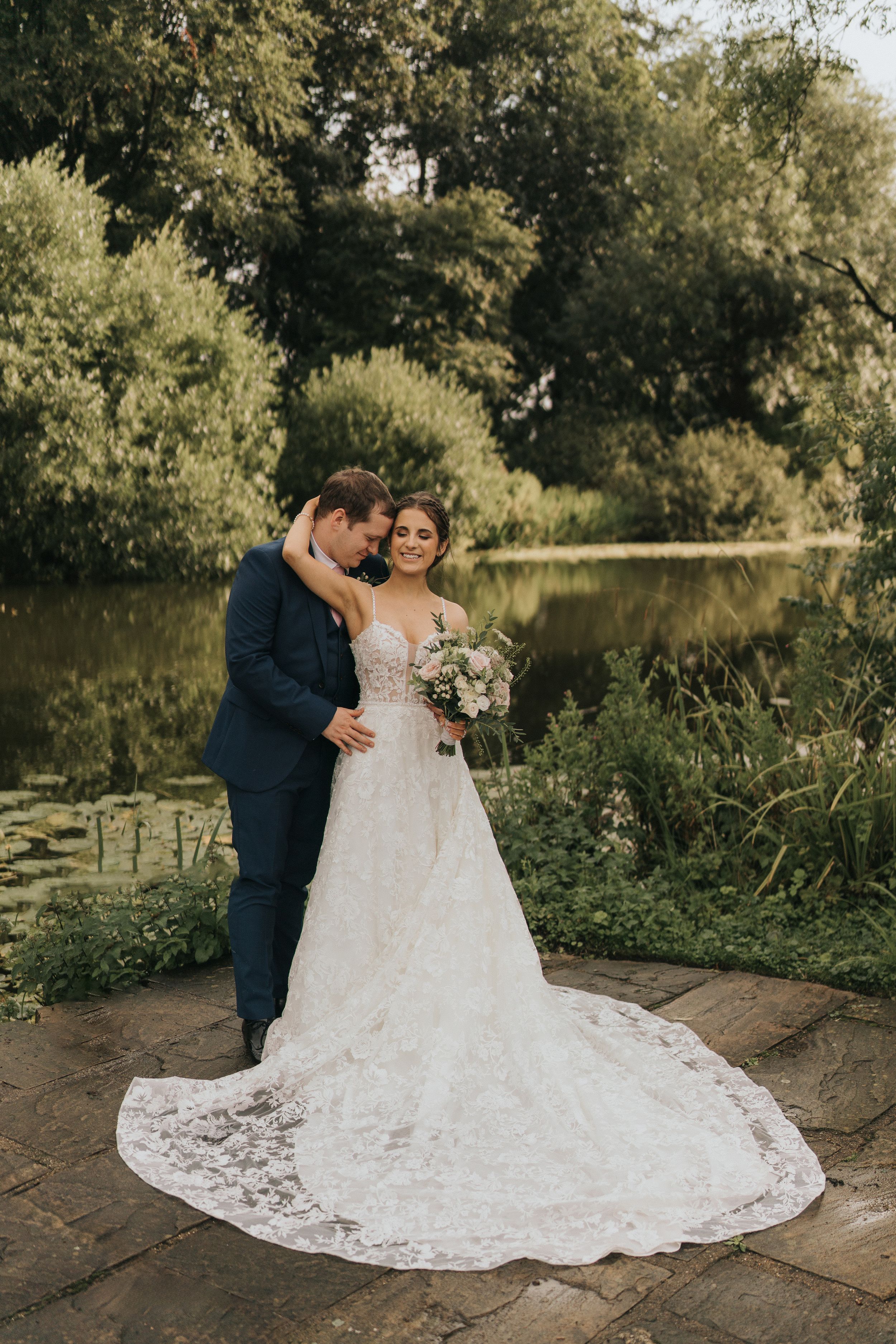A couple shares an intimate moment in their wedding attire by a tranquil pond surrounded by lush greenery at Cave Castle.