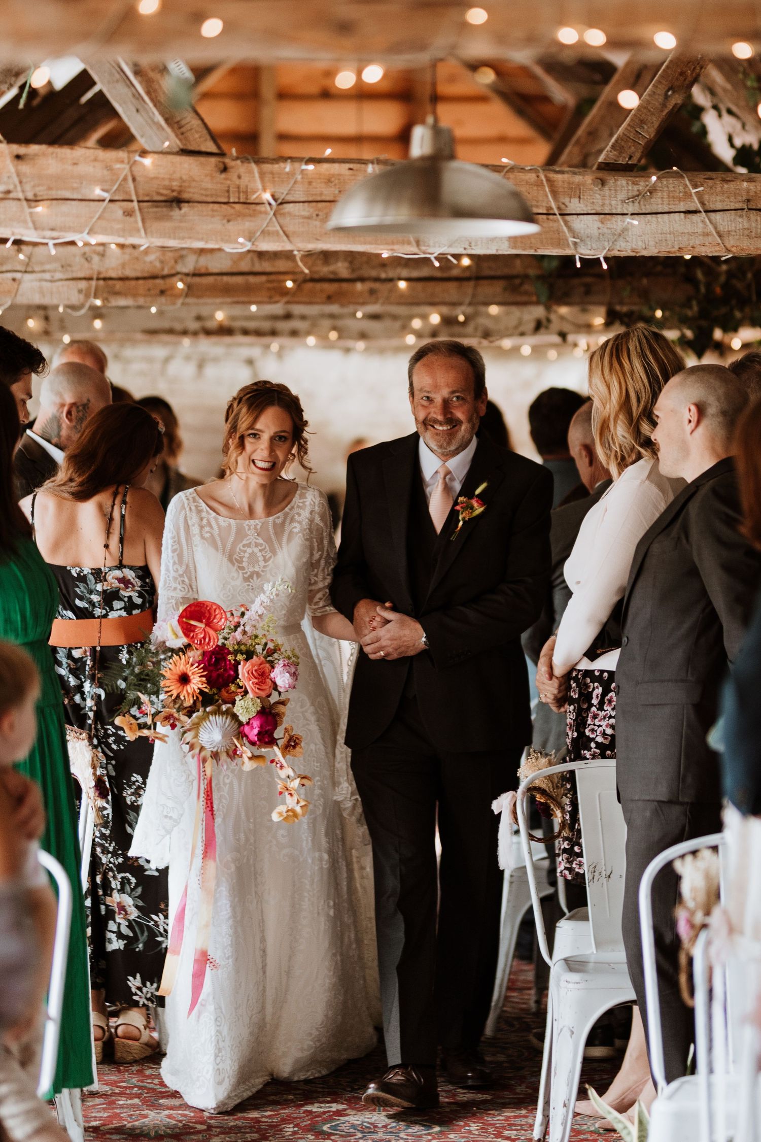 A bride in a lace gown walks down the aisle at a rustic barn wedding ceremony holding a vibrant bouquet.
