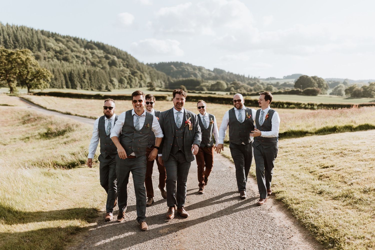 A group of men in matching gray vests and dark pants walk together along a rural path surrounded by green countryside.