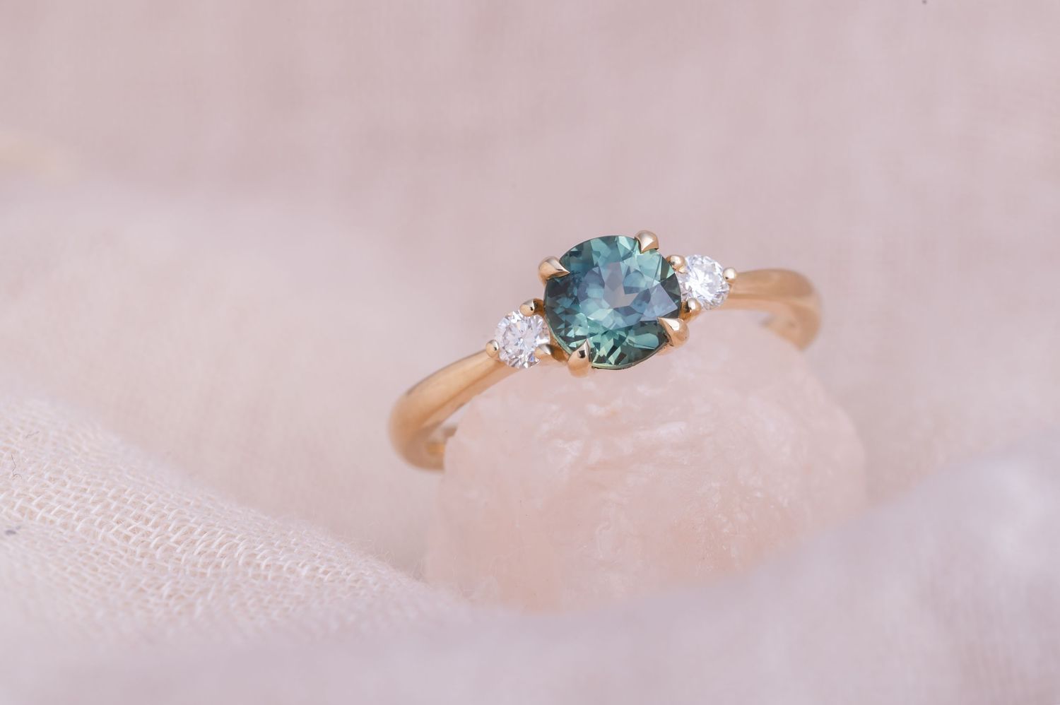 Close-up of an elegant teal gemstone ring with small diamond accents on pink fabric.