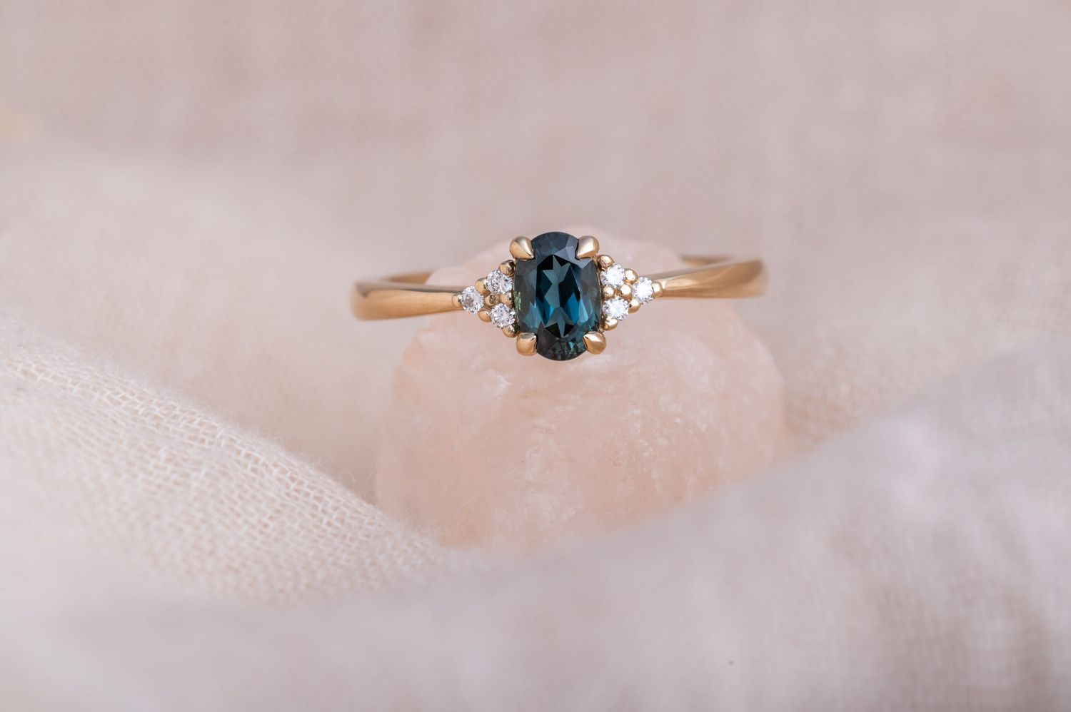 Teal sapphire ring with diamond side stones photographed on light fabric.