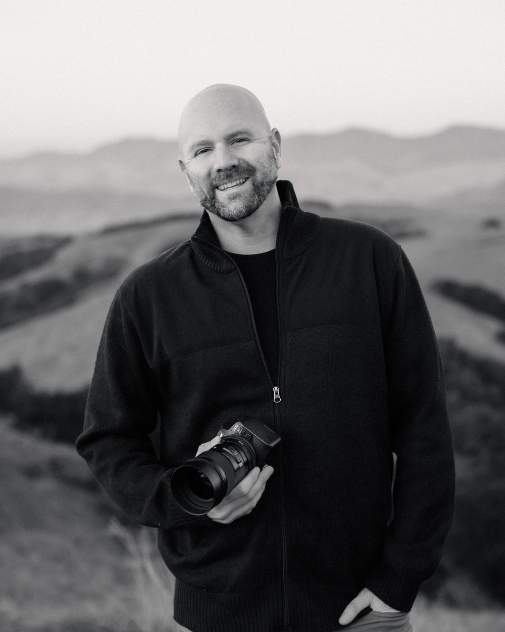 Portrait of a photographer holding a camera against rolling hills and mountains in monochrome.