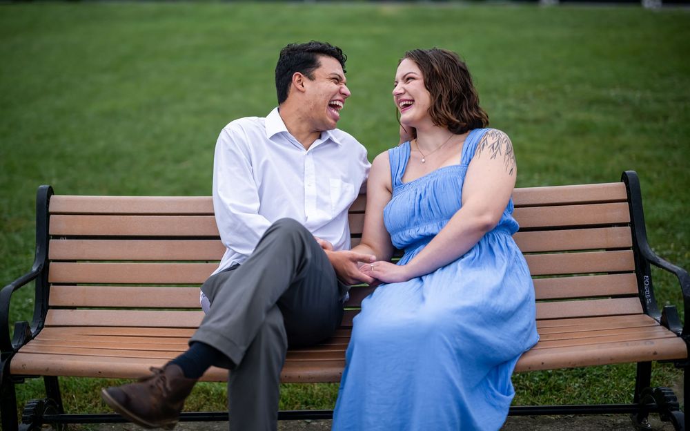 Romantic couple sit together and laugh on a wooden park bench in matching grey and blue outfits on a sunny day, in Baltimore.