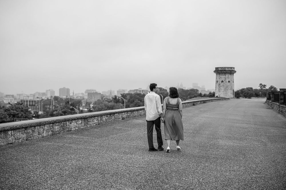 A romantic couple in black and white stand in distance at the edge of a wall, looking at a scenic view, in baltimore.