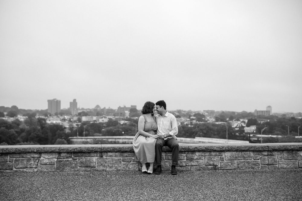 A couple shares an intimate moment sitting on a stone wall overlooking a city skyline in black and white, In baltimore