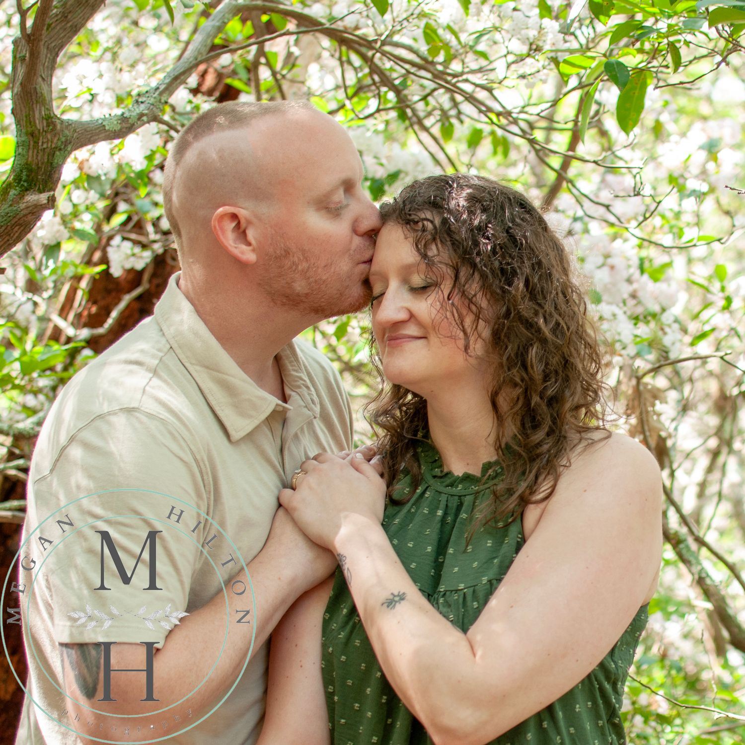 A romantic couple share a gentle moment embracing under blooming tree branches in a sunny nature setting.