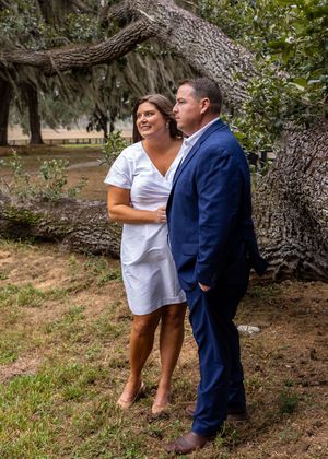 Couple in formal wear standing by large tree trunk in outdoor setting.