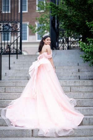 Person in flowing pink gown poses dramatically on stone steps near iron gate.