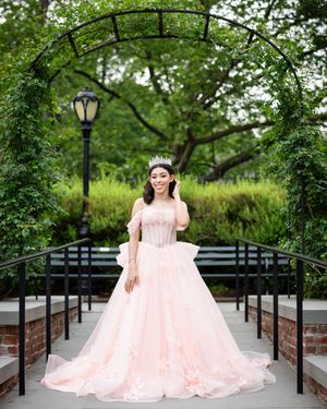 Young woman in flowing pink gown poses under garden archway with vintage lamppost.
