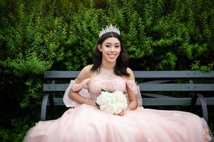 Person in pink ballgown and tiara sits on garden bench holding white bouquet.