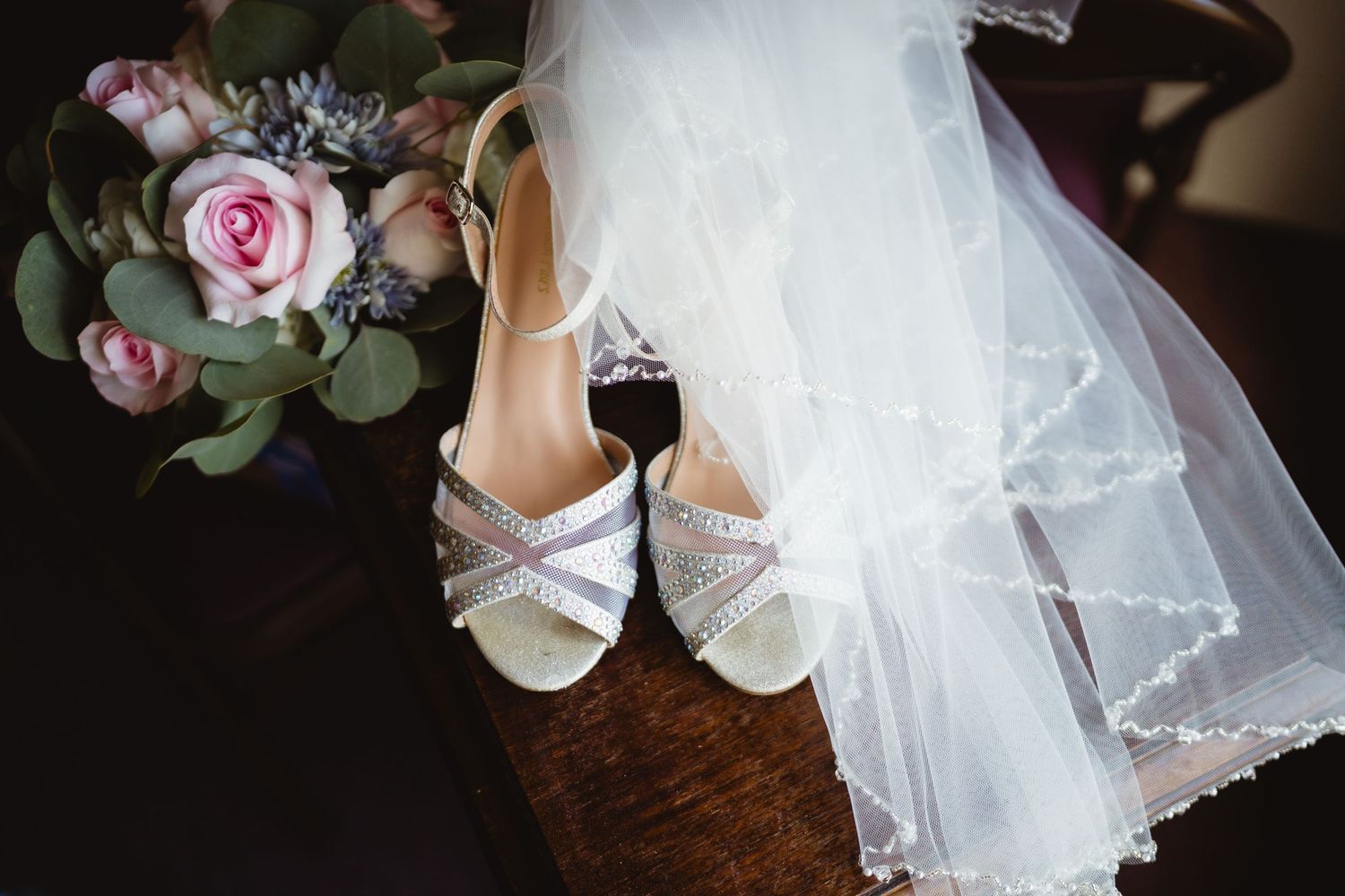Wedding details display with white bridal shoes, delicate veil, and pink rose bouquet arranged on wooden surface.