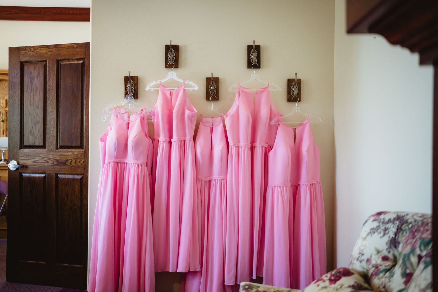 Pink bridesmaid dresses hang neatly in a row against cream colored walls of an indoor space.