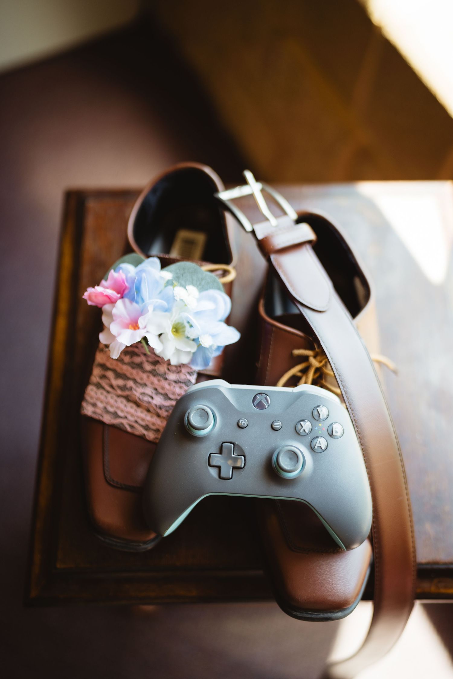 Xbox controller and leather belt rest on wooden surface next to delicate floral arrangement.