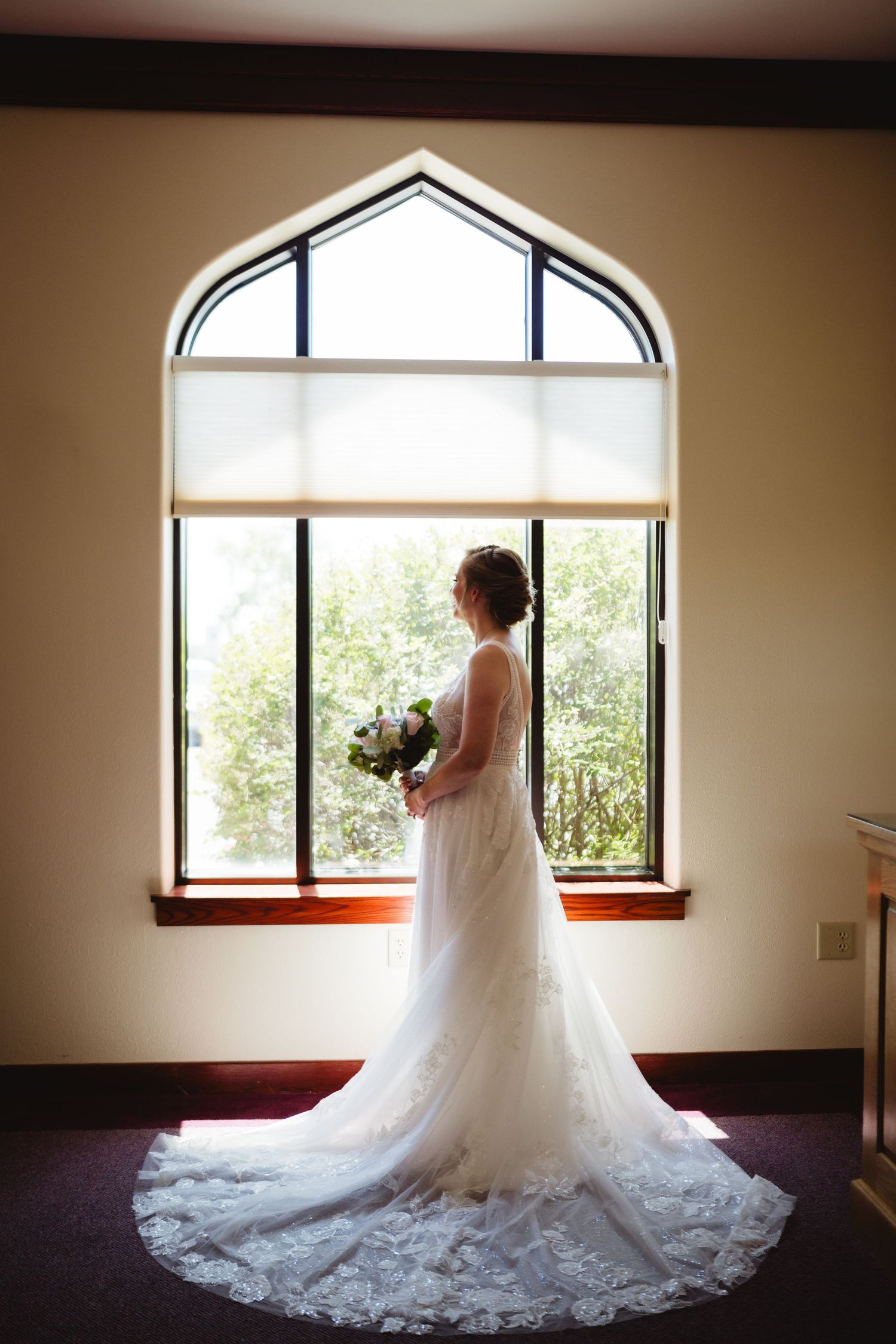 Silhouetted figure in flowing lace wedding dress stands by arched church window with bouquet in natural light.