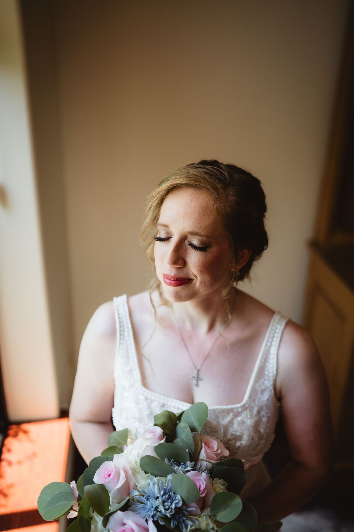 A bride in white dress holds pastel bouquet with eucalyptus while looking down near window in soft natural light.