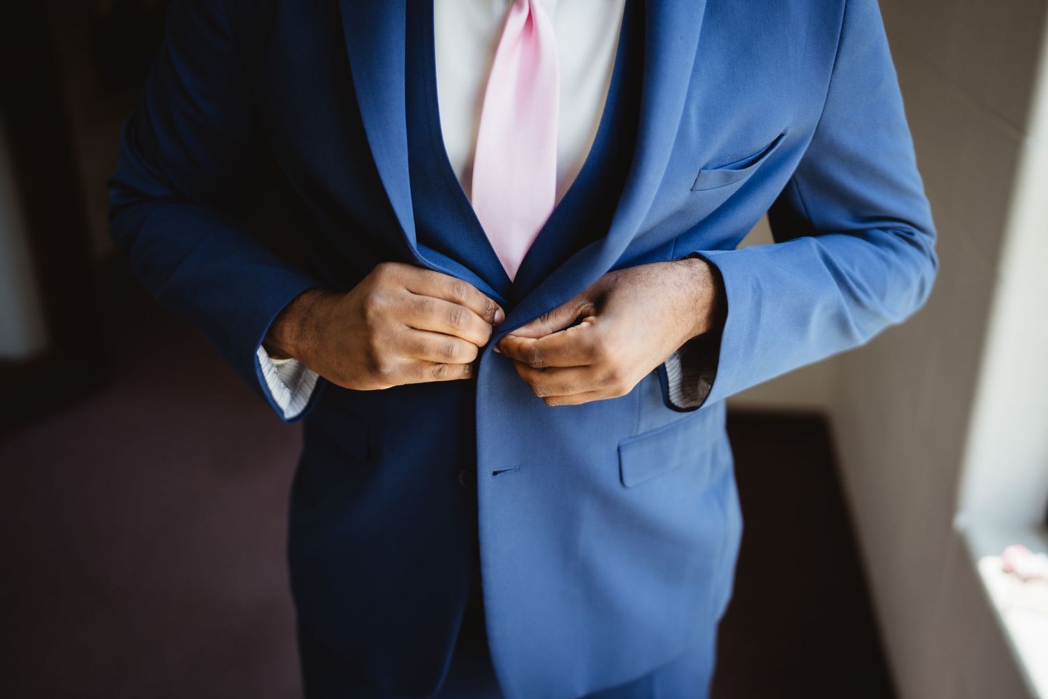 A person wearing a bright blue suit jacket and pink tie buttons their coat in a shadowy room.