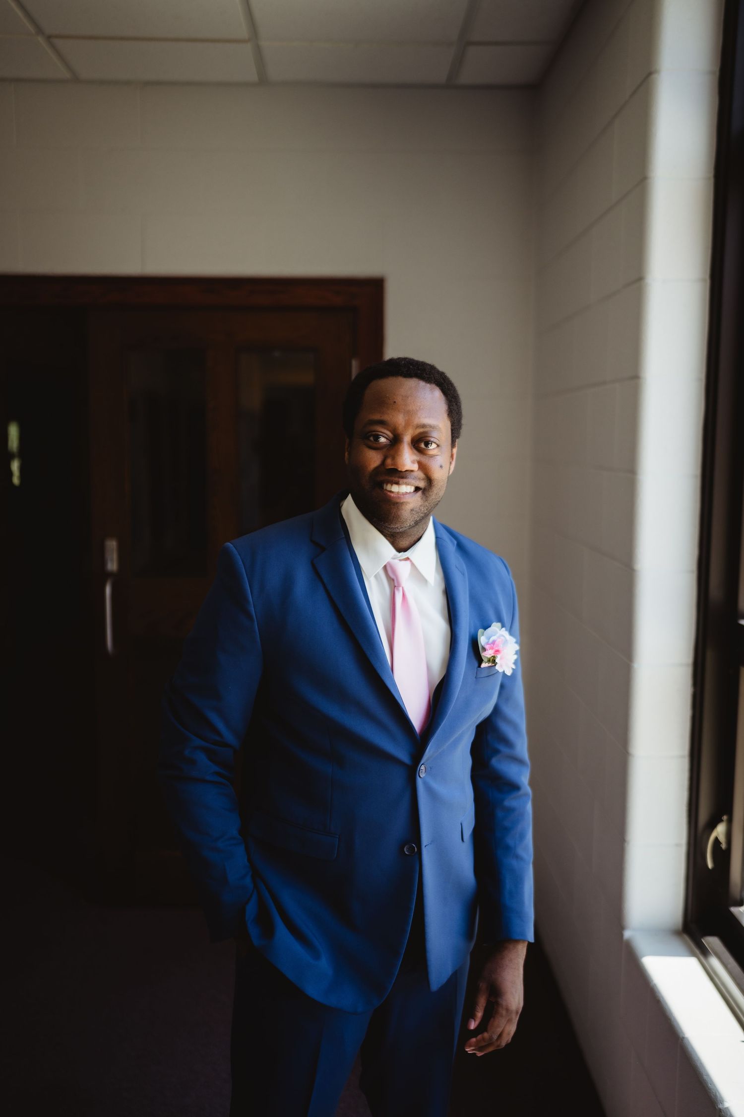 A groomsman wearing a royal blue suit and pink tie poses near a window in an elegant indoor setting.